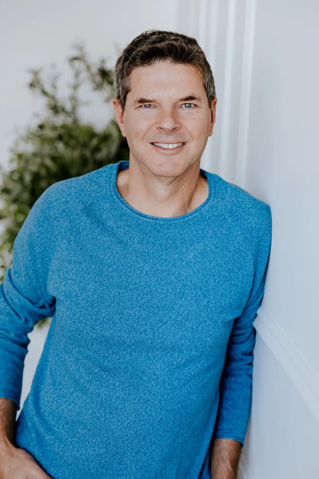 Portrait de Luc Turell, souriant, adossé à un mur clair près d'un feuillage vert.